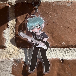 Könnte beinhalten: Ein klarer Acryl-Schlüsselanhänger mit einem Anime-Charakter mit türkisfarbenen Haaren, der eine schwarz-weiße Gitarre hält. Der Charakter trägt ein schwarzes Outfit. Der Schlüsselanhänger ist an einem silbernen Schlüsselring befestigt.