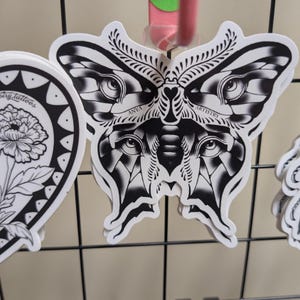 May include: Black and white butterfly sticker with intricate details, featuring eye-like patterns and the words "ANYA ARTISTRY". The sticker is displayed on a black wire grid, showcasing a tattoo-inspired design.
