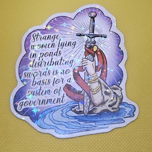 May include: A sticker with a purple and blue design featuring a sword held by a hand emerging from water. The text reads "Strange women lying in ponds distributing swords is no basis for a system of government."