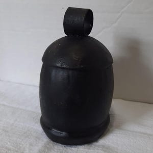 May include: A black, bell-shaped container with a rounded top and a small, cylindrical handle. The container has a smooth, matte finish and sits on a slightly wider base. The background is a plain, off-white surface.