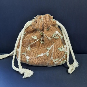 May include: A small, drawstring pouch in shades of brown, with a delicate floral overlay. The bag features a woven texture and is accented with cream-colored rope drawstrings and tassels. The floral design includes white and green accents.