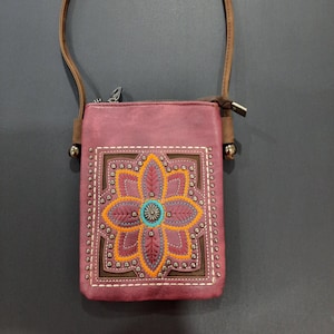 May include: A burgundy crossbody bag with a floral design. The bag features a stitched flower in orange, blue, and burgundy, framed by a brown square with silver studs. It has a brown strap and a zipper closure.