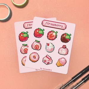 May include: Two pink cards with pixelated strawberry-themed illustrations. Each card features a variety of strawberry-related images, including strawberries, desserts, and the word "Strawberry" at the top. The cards are on a pink surface with pens and tape.