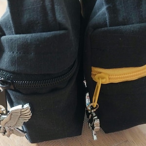 May include: Two black fabric pouches with rectangular structures. The left pouch features a black zipper with a silver-toned eagle charm. The right pouch has a yellow zipper with a small silver-toned charm attached to the pull tab.
