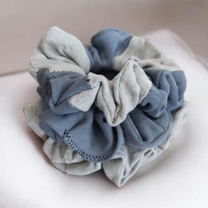 Puede incluir: Una scrunchie hecha a mano en tonos grises y azules. La tela está fruncida y cosida, creando un efecto texturizado y en capas. La scrunchie está diseñada para accesorios para el cabello.