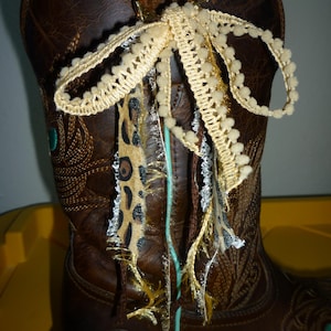 May include: A brown leather western boot featuring a turquoise piping accent and an Ariat logo embossed on the side. A decorative bow made of cream-colored pom-pom trim, leopard print fabric strips, and frayed metallic gold ribbon is tied to the pull tab.