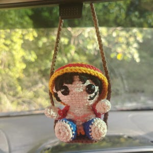 Peut inclure: Poupée Luffy crochetée suspendue au rétroviseur d'une voiture. La poupée porte un chapeau jaune et rouge, des cheveux noirs et une chemise rouge. Elle est assise sur une balançoire crochetée avec un short bleu et blanc.