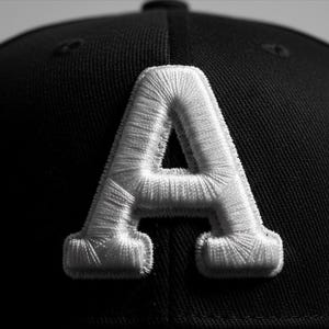 以下が含まれることがあります： 前面に大きな白い刺繍文字「A」が付いた黒い野球帽。 「A」は、テクスチャ加工された隆起したデザインでステッチされています。 キャップは濃い色の生地でできており、詳細な刺繍に焦点が当てられています。