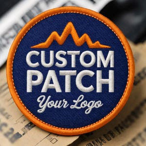 Op de afbeelding: Een ronde, marineblauwe custom patch met een oranje rand. De patch heeft een oranje bergketenontwerp boven de witte tekst "CUSTOM PATCH" en de cursieve tekst "Your Logo".