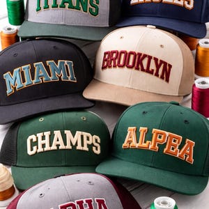 Multiple snapback hats featuring different color combinations of 3D puff varsity embroidery font styles
