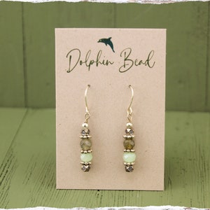 May include: Gold dangle earrings with green and brown beads. The earrings are on a card that says "Dolphin Bead".