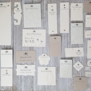 May include: Assortment of blank, cream-colored and brown paper tags with the text "Hand Crafted Designs" and various designs. Some tags have holes for hanging, others have a decorative edge. The tags are arranged on a wooden surface.