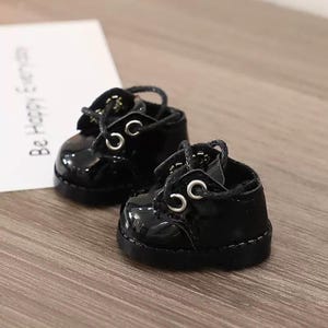 May include: A pair of miniature black patent leather shoes with laces and silver eyelets. The shoes are glossy and sit on a wooden surface. A white card with the text "Be Happy Everyday" is in the background.