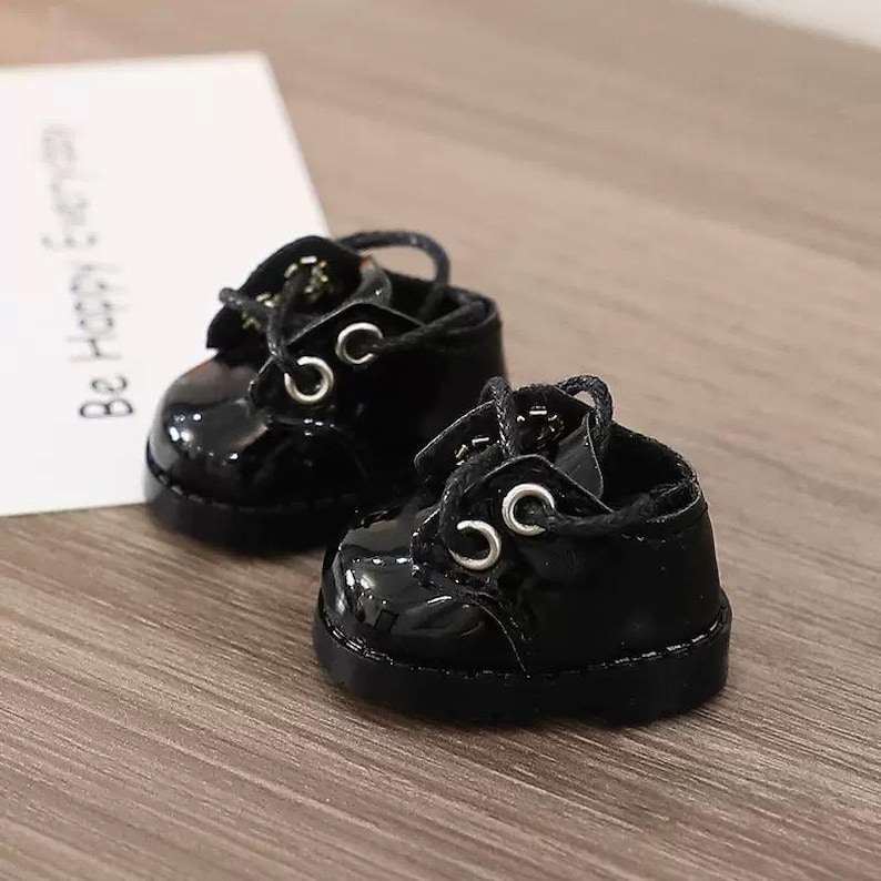 May include: A pair of miniature black patent leather shoes with laces and silver eyelets. The shoes are glossy and sit on a wooden surface. A white card with the text "Be Happy Everyday" is in the background.