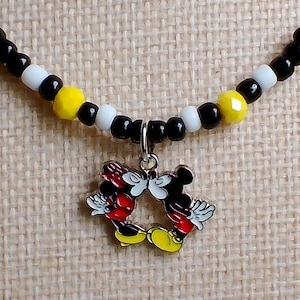 May include: A beaded necklace featuring a silver-toned metal charm of two cartoon mice kissing. The necklace is strung with a repeating pattern of small black, white, and faceted yellow beads. The charm depicts two figures with red shorts and yellow shoes.