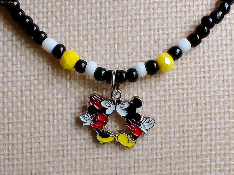 May include: A beaded necklace featuring a silver-toned metal charm of two cartoon mice kissing. The necklace is strung with a repeating pattern of small black, white, and faceted yellow beads. The charm depicts two figures with red shorts and yellow shoes.