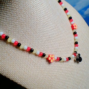 May include: A beaded necklace with red, black, white, and yellow beads. The necklace features two red flower-shaped charms with yellow centers and a small black charm at the clasp. The necklace is displayed on a beige surface.