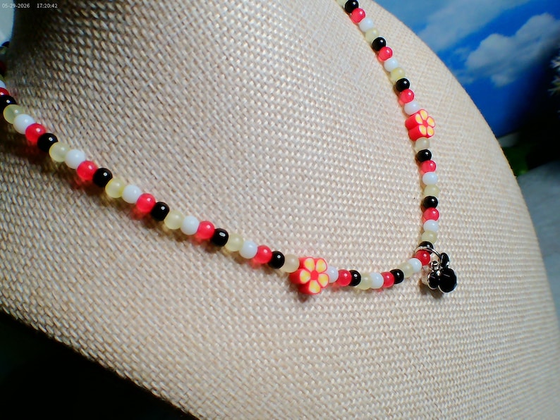 May include: A beaded necklace with red, black, white, and yellow beads. The necklace features two red flower-shaped charms with yellow centers and a small black charm at the clasp. The necklace is displayed on a beige surface.