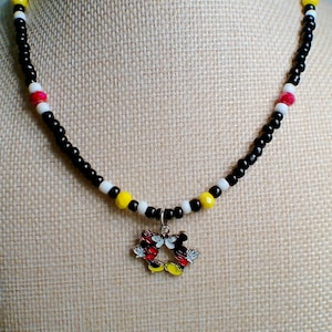 May include: A beaded necklace featuring a central metal charm of two cartoon mice kissing. The necklace is constructed with small black, white, yellow, and red beads arranged in a repeating pattern along the entire length of the circular strand.
