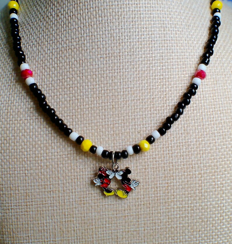 May include: A beaded necklace featuring a central metal charm of two cartoon mice kissing. The necklace is constructed with small black, white, yellow, and red beads arranged in a repeating pattern along the entire length of the circular strand.