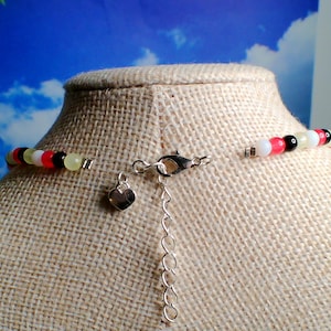 May include: A beaded necklace with a silver clasp and heart-shaped charm. The necklace features a colorful strand of beads in white, pink, black, yellow, and red. The necklace is displayed on a mannequin.