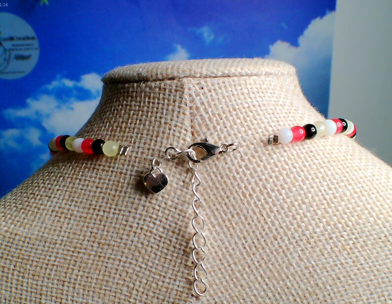 May include: A beaded necklace with a silver clasp and heart-shaped charm. The necklace features a colorful strand of beads in white, pink, black, yellow, and red. The necklace is displayed on a mannequin.