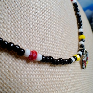 May include: A beaded necklace featuring a repeating pattern of small black and white spherical beads, accented with occasional red and yellow faceted beads. A small silver-toned metal charm with red, yellow, and black enamel details hangs from the center.