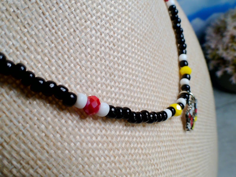 May include: A beaded necklace featuring a repeating pattern of small black and white spherical beads, accented with occasional red and yellow faceted beads. A small silver-toned metal charm with red, yellow, and black enamel details hangs from the center.