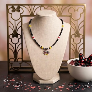 May include: A beaded necklace featuring small black beads interspersed with yellow, white, and red accent beads. A central charm shaped like a flower with colorful petals hangs from the necklace, which is displayed on a beige jewelry bust.