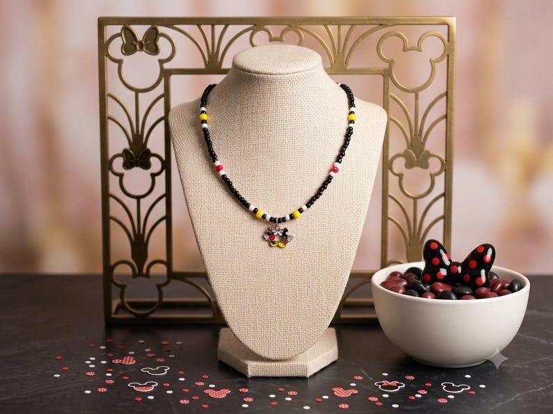 May include: A beaded necklace featuring small black beads interspersed with yellow, white, and red accent beads. A central charm shaped like a flower with colorful petals hangs from the necklace, which is displayed on a beige jewelry bust.