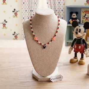 May include: A beaded necklace displayed on a beige bust. The necklace features a pattern of white, black, and red beads, with orange flower accents and a Mickey Mouse pendant. The background includes Mickey Mouse decor and a handmade tag.