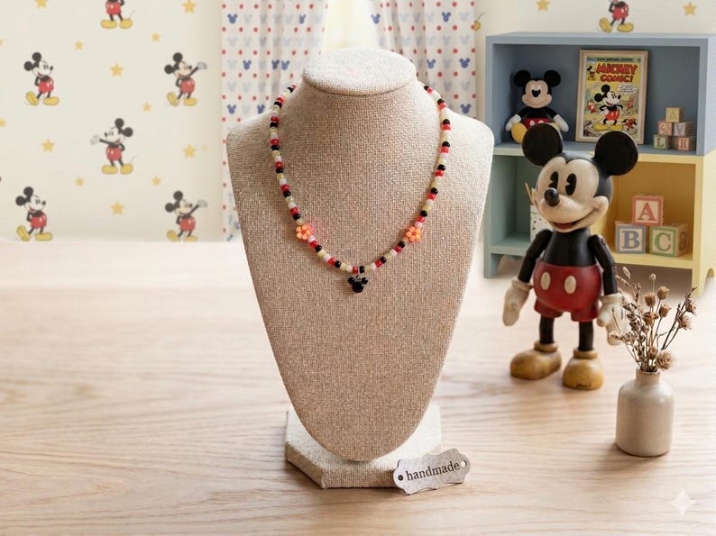 May include: A beaded necklace displayed on a beige bust. The necklace features a pattern of white, black, and red beads, with orange flower accents and a Mickey Mouse pendant. The background includes Mickey Mouse decor and a handmade tag.