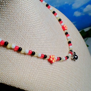 May include: A beaded necklace with red, white, black, and yellow beads. The necklace features small, red flower-shaped beads. The necklace is displayed on a mannequin.