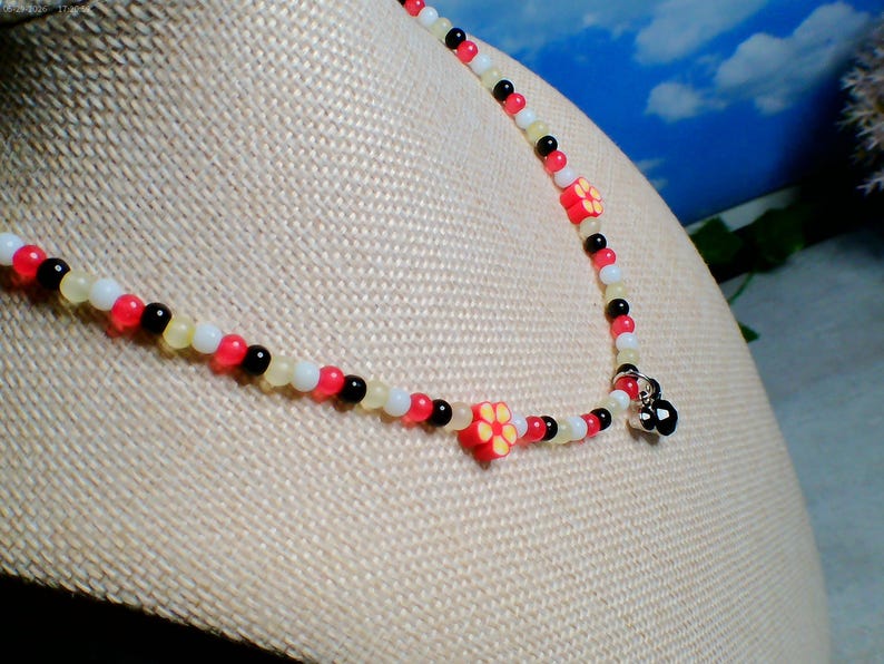 May include: A beaded necklace with red, white, black, and yellow beads. The necklace features small, red flower-shaped beads. The necklace is displayed on a mannequin.