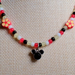 May include: A beaded necklace with a black Mickey Mouse pendant. The necklace features red, white, black, and yellow beads, with two flower-shaped beads. The necklace is displayed on a beige fabric background.