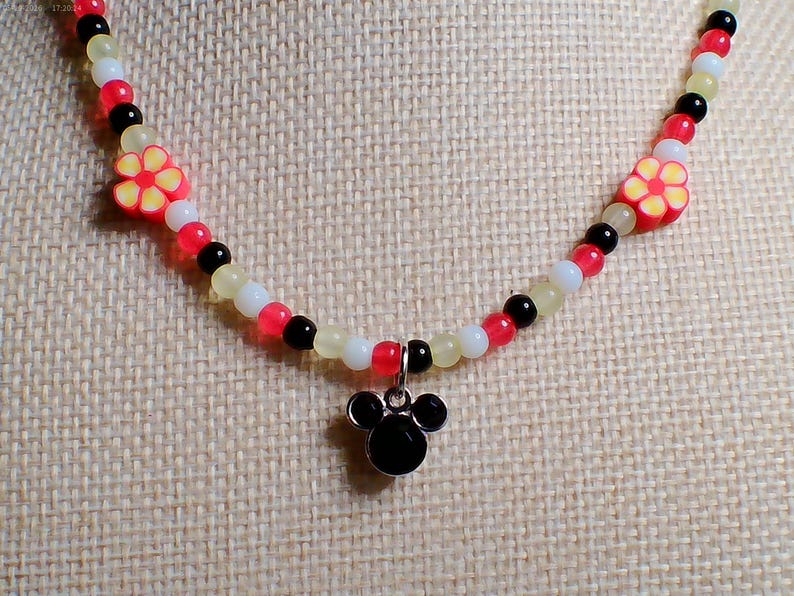 May include: A beaded necklace with a black Mickey Mouse pendant. The necklace features red, white, black, and yellow beads, with two flower-shaped beads. The necklace is displayed on a beige fabric background.