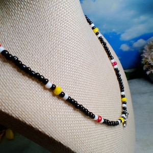 May include: A beaded necklace featuring a repeating pattern of small black seed beads interspersed with white, bright yellow, and faceted red beads. The necklace is strung on a flexible cord and includes a small silver-toned metal charm at the center.