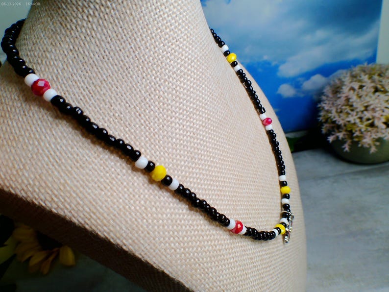 May include: A beaded necklace featuring a repeating pattern of small black seed beads interspersed with white, bright yellow, and faceted red beads. The necklace is strung on a flexible cord and includes a small silver-toned metal charm at the center.