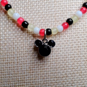 May include: A beaded necklace featuring a black Mickey Mouse pendant. The necklace has a strand of small beads in red, black, white, and yellow. The pendant is a black, stylized silhouette of Mickey Mouse's head.