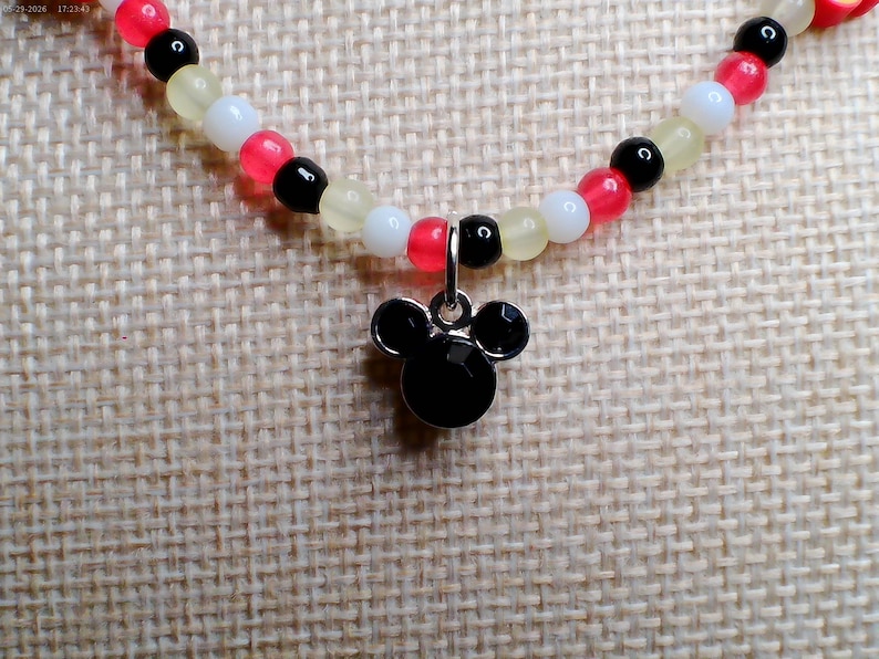 May include: A beaded necklace featuring a black Mickey Mouse pendant. The necklace has a strand of small beads in red, black, white, and yellow. The pendant is a black, stylized silhouette of Mickey Mouse's head.