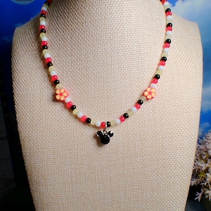 May include: A beaded necklace with a black, red, white, and yellow color scheme. The necklace features two flower accents and a black Mickey Mouse-shaped pendant. The necklace is displayed on a mannequin.