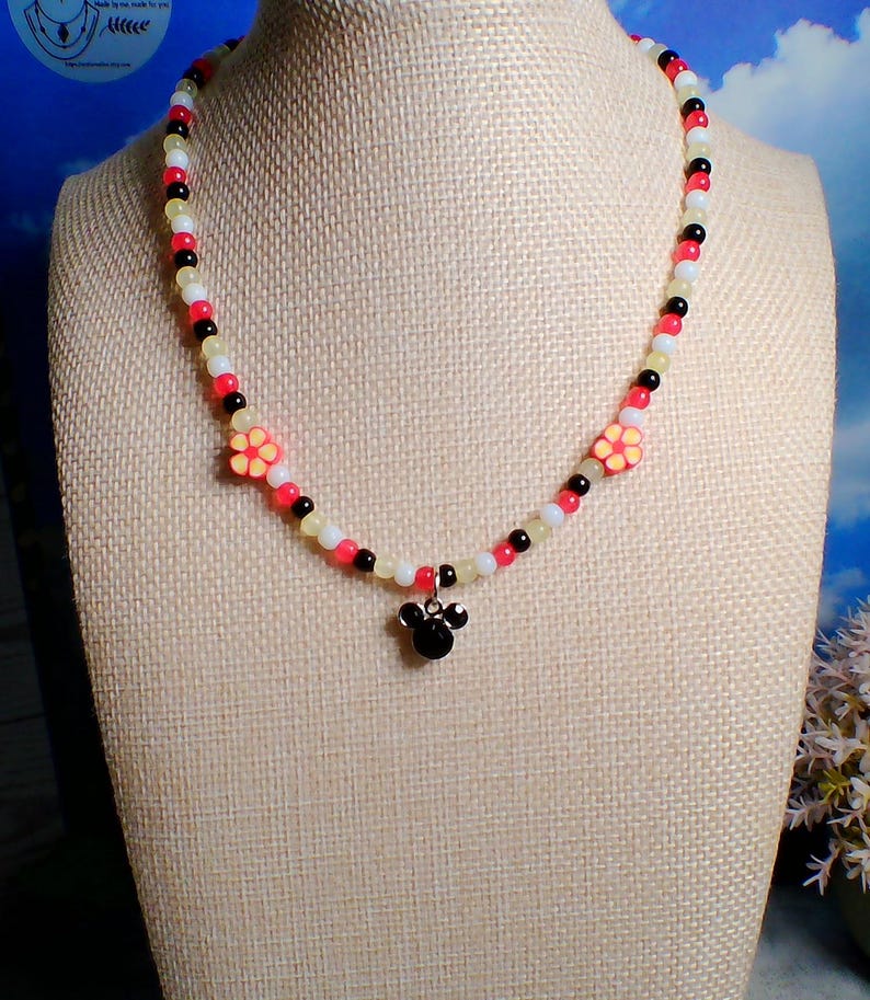 May include: A beaded necklace with a black, red, white, and yellow color scheme. The necklace features two flower accents and a black Mickey Mouse-shaped pendant. The necklace is displayed on a mannequin.