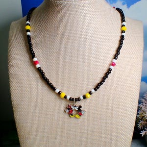 May include: A beaded necklace featuring a central charm of two cartoon mouse characters joined together. The necklace is strung with small black beads, interspersed with white, yellow, and red accent beads in a repeating color pattern.