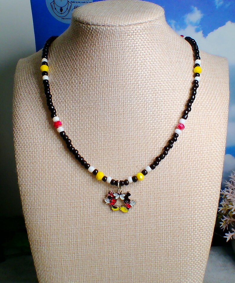 May include: A beaded necklace featuring a central charm of two cartoon mouse characters joined together. The necklace is strung with small black beads, interspersed with white, yellow, and red accent beads in a repeating color pattern.