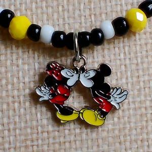May include: A beaded necklace featuring a silver-toned metal charm of two cartoon mice kissing. The necklace is strung with alternating round black, white, and faceted yellow beads. The charm depicts two figures with red clothing and yellow shoes.