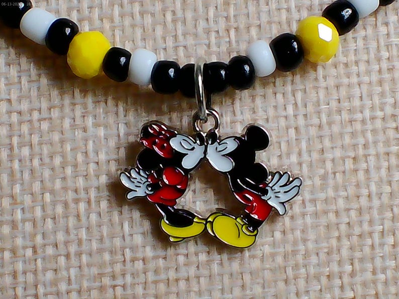 May include: A beaded necklace featuring a silver-toned metal charm of two cartoon mice kissing. The necklace is strung with alternating round black, white, and faceted yellow beads. The charm depicts two figures with red clothing and yellow shoes.