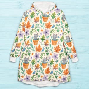 Può includere: Una coperta indossabile color crema con un motivo stravagante di gatti grigi in vaso, foglie di acero arancioni, uccelli blu e fiori viola. La coperta ha un cappuccio e polsini foderati in pile bianco e una grande tasca frontale.
