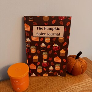 Op de afbeelding: Een herfststillevens met een notitieboek "The Pumpkin Spice Journal", een oranje kaars en een decoratieve pompoen. Het notitieboek heeft een bruine kaft met illustraties van herfstlekkernijen.
