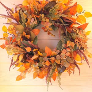 May include: A circular autumn wreath featuring a dense arrangement of faux foliage in shades of orange, yellow, and deep brown. The wreath includes various leaf shapes, textured grasses, small berry clusters, and dark green magnolia-style leaves.
