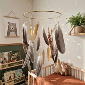 Peut inclure: Mobile pour bébé avec un anneau doré et des plumes suspendues dans des tons marron, gris et or. Les plumes sont suspendues par de fins fils, avec des accents de cristal transparent. Le mobile est au-dessus d'un lit bébé en bois.